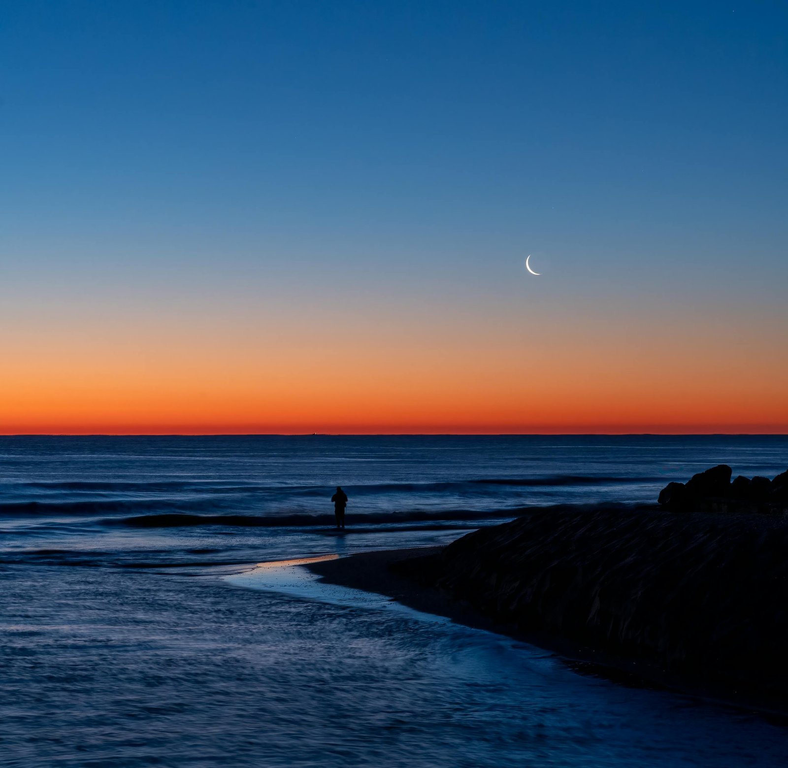 A tranquil twilight scene of a beach in Valencia, Spain, with a crescent moon.
